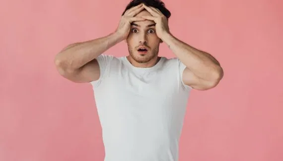 Imagem mostra um homem de camiseta branca com a mão na cabeça desesoerado.