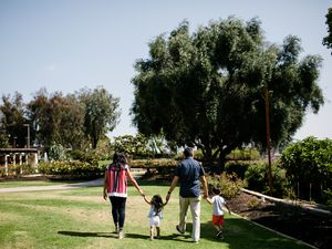 Family of four walking in grass through a garden facing away from camera. A brunette woman is one the left, hold hands with a young girl. The girl is also holding hands with a brunette man who is holding hands with a young boy. 