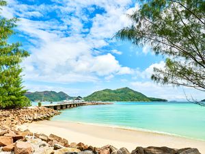 Pier jetty next to a beach on Praslin island, Seychelles. The water is a sea foam green and there are green mountainous islands across the water