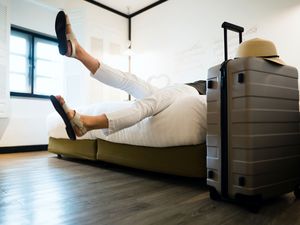 Young female traveller relaxing on bed in a hotel room, with a suitcase and straw hat by the bed