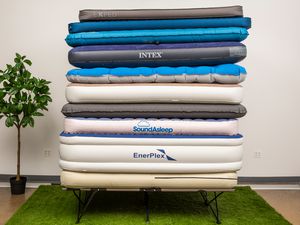 Stack of camping air mattresses we recommend on a green rug next to a plant 