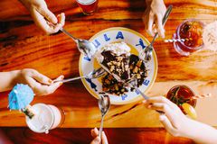 Five hands holding spoons going to eat a slice of hul pie covered in chocolate sauce on a plate that says "hula pie. There are four drinks on the table as well