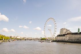 Large white ferris wheel on the right side of a wide river with a pale blue sky and several clouds