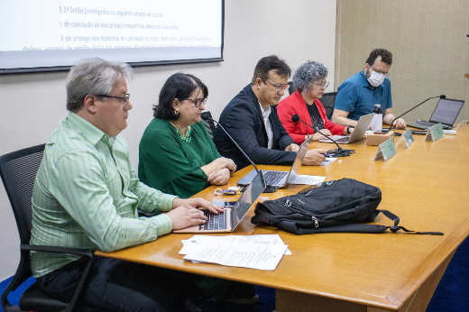 Imagem: mesa retangular onde cinco pessoas estão sentadas, participando de uma reunião ou evento. Todas têm notebooks ou documentos à frente. Da esquerda para a direita: um homem de cabelo grisalho com camisa listrada verde, uma mulher de óculos com roupa verde, um homem de óculos com blazer preto, uma mulher de cabelo grisalho e casaco vermelho, e um homem com máscara facial azul e camiseta verde. No fundo, há um projetor exibindo texto na parede. Sobre a mesa, há uma mochila e papéis espalhados.