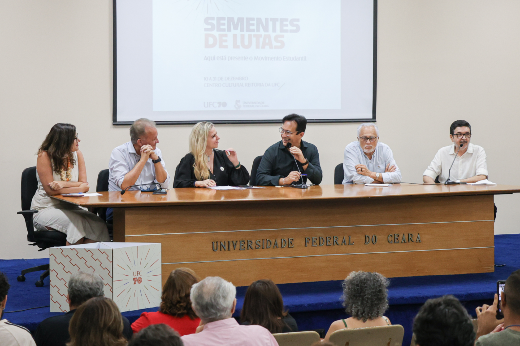 Imagem: A imagem retrata uma mesa de debate composta por seis pessoas, em um auditório. Os participantes estão sentados atrás de uma mesa longa de madeira com a inscrição "Universidade Federal do Ceará" na frente. O evento parece ser formal e acadêmico.  Ao fundo, uma tela de projeção exibe o título "Sementes de Lutas", indicando o tema da discussão. A frase "Aqui está presente o Movimento Estudantil" também aparece no slide projetado, reforçando a relação com pautas estudantis.  Cada participante está vestindo roupas formais ou semi-formais, e todos estão posicionados em frente a microfones. Eles parecem atentos e engajados, como se estivessem discutindo ou ouvindo a fala de um colega.  No público, várias pessoas estão sentadas em cadeiras, algumas com cabelos grisalhos, sugerindo diversidade de idades. Uma pessoa na plateia segura um celular, possivelmente registrando o momento. O ambiente é claro e bem iluminado, típico de um evento acadêmico em um auditório.