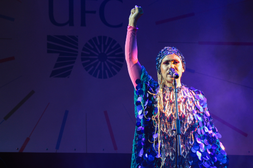 Imagem: Uma mulher está no palco sob luzes coloridas, segurando um dos braços erguidos com o punho fechado. Ela está cantando ou falando em um microfone e usa uma roupa reluzente decorada com paetês azulados e brilhantes, além de uma touca que cobre a cabeça. Ao fundo, há um painel grande com o texto "UFC 70" e linhas coloridas que parecem marcar o tempo, como em um relógio estilizado.