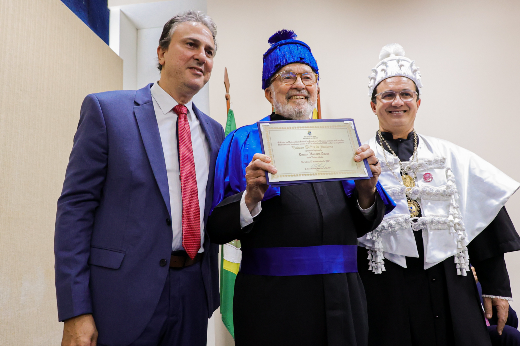 Imagem: Três homens estão posando para uma foto em um evento formal. O homem ao centro está vestido com uma beca azul e preta, usando um chapéu azul, e segura um diploma ou certificado sorrindo. À esquerda, há um homem de terno azul e gravata vermelha. À direita, outro homem veste uma beca branca e preta, com um chapéu branco ornamentado, também sorrindo. Ao fundo, há uma bandeira parcialmente visível. A cena indica um evento acadêmico ou de homenagem oficial.