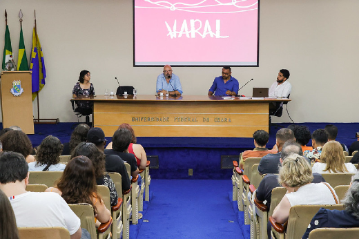 Imagem: A imagem mostra uma mesa redonda em um auditório da Universidade Federal do Ceará (UFC). Quatro pessoas estão sentadas atrás de uma mesa no palco, participando de um debate. Acima delas, há um telão exibindo o título "Varal" em destaque. A plateia está composta por diversas pessoas sentadas, acompanhando o evento. No fundo à esquerda, há bandeiras em suportes.