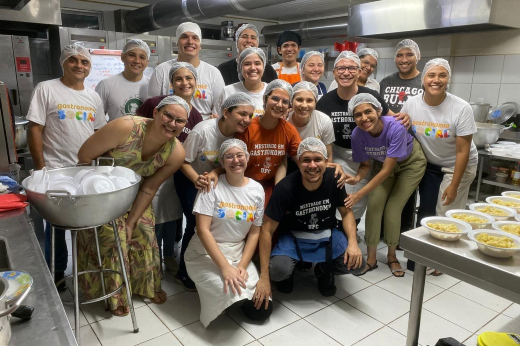 Imagem: grupo de cerca de 20 pessoas reunidas em uma cozinha industrial. Todos estão sorrindo e usam toucas de proteção, alguns também vestem camisetas que indicam "Gastronomia Social" e outras mensagens relacionadas à culinária. Ao fundo, há equipamentos de cozinha, como fogões e bancadas, e, na frente, uma mesa com pratos de comida prontos. O ambiente parece acolhedor e de trabalho em equipe.