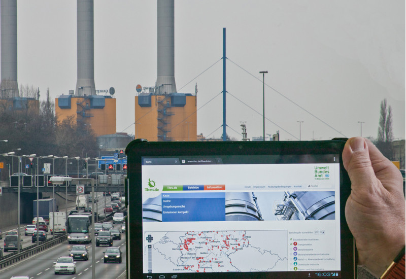 eine Hand hält ein Tablet mit dem Webangebot thru.de vor ein Kraftwerk hinter einer Stadtautobahn