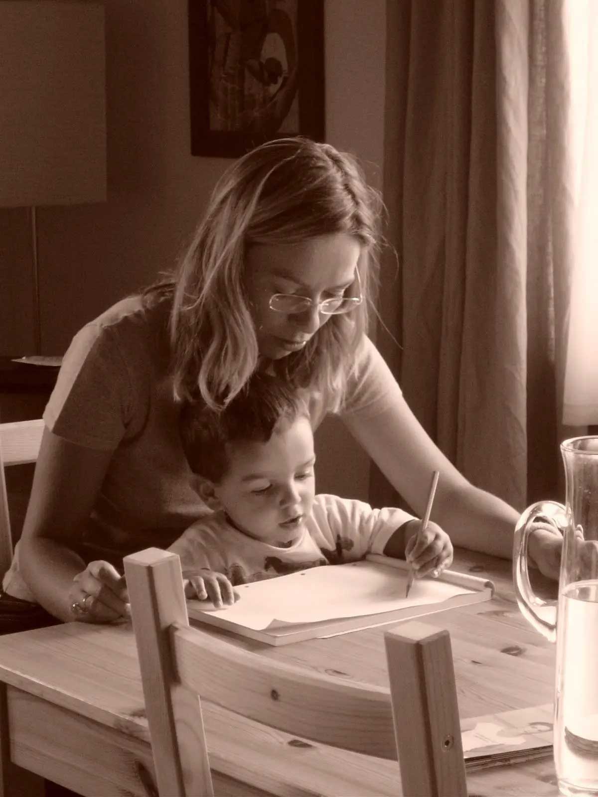 At a table, Laura sits with her young son on her lap while he draws and she looks on over his head