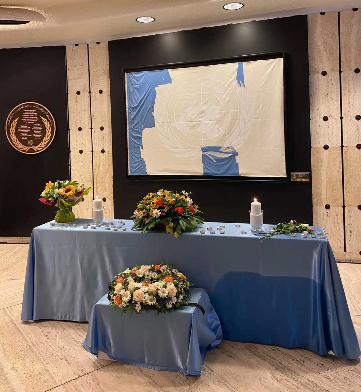 tables with blue cloth, flower bouquets, and candles. A torn UN flag is seen behind.