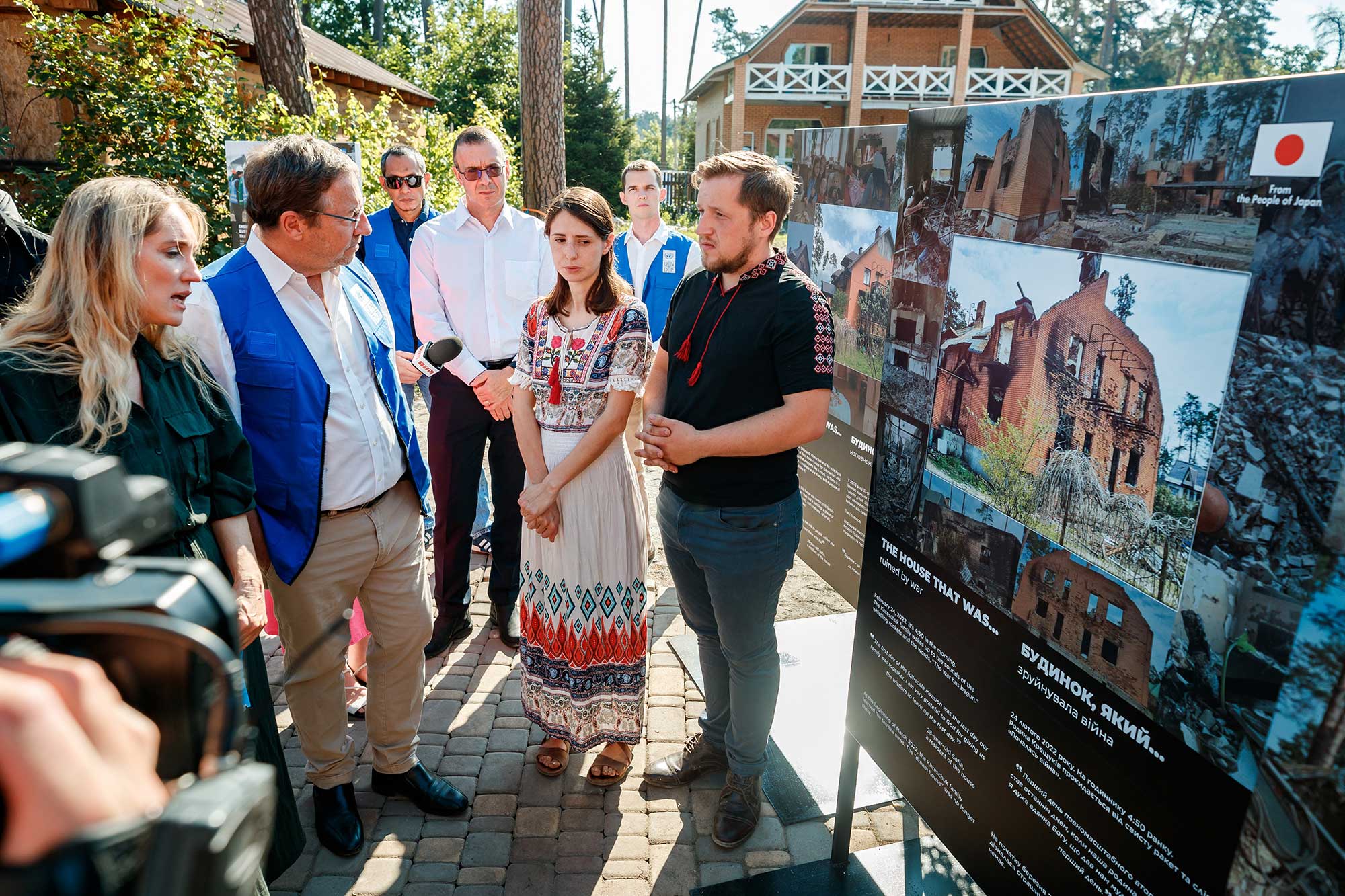 Achim speaking with a woman and a man who are dressed in traditional Ukranian clothes. They are all surrounded by reporters with microphones and stand next to a poster.