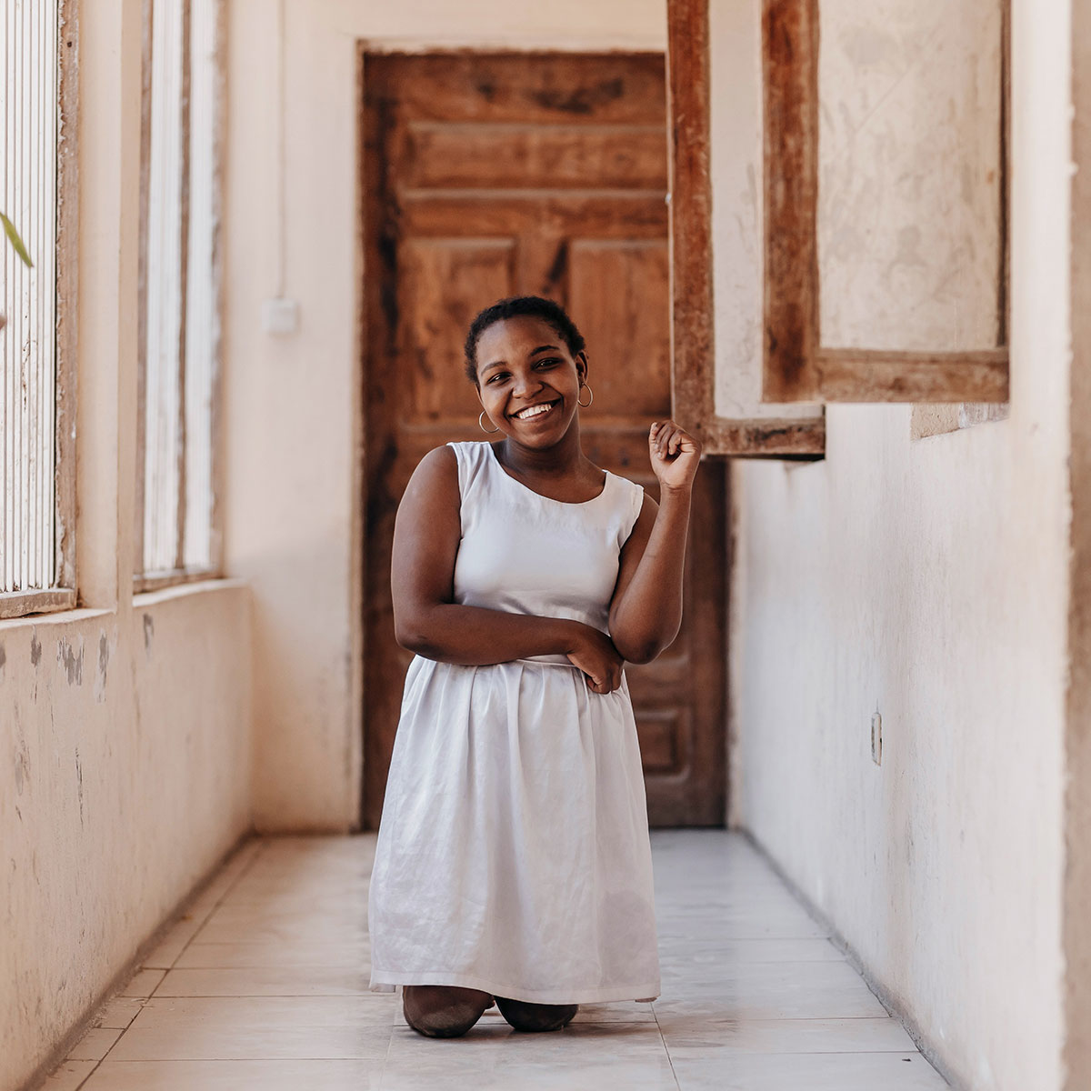 A young woman in a white dress kneels in a light-filled hallway, looking straight ahead with a big smile on her face
