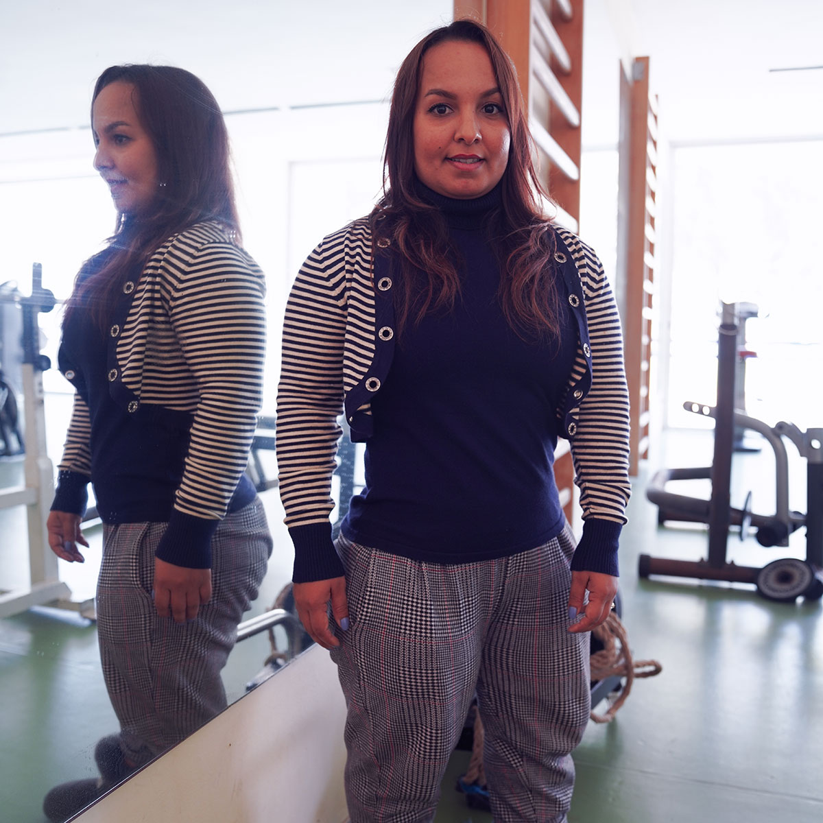 Hayat stands in a fitness centre, looking straight ahead and smiling, reflected in a large mirror to her right