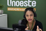 Uma mulher com fones de ouvido está sentada em frente a um computador, aparentemente engajada em uma videochamada ou gravação. Ela usa uma blusa preta e um crachá de identificação verde da Unoeste. A mulher gesticula com as mãos enquanto fala, segurando uma caneta na mão direita. Ao fundo, uma parede verde exibe o logotipo da Unoeste e a frase 