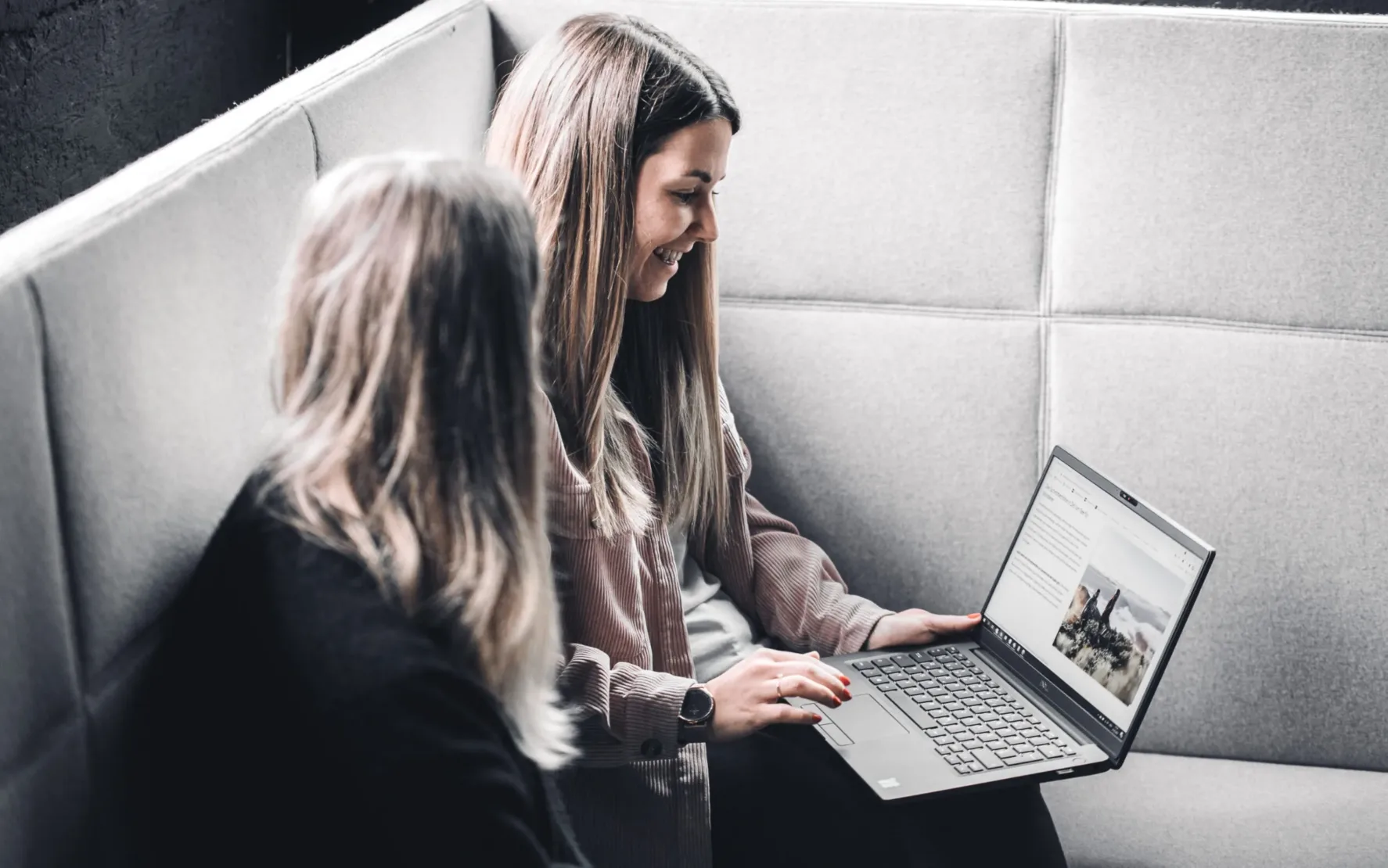 Zwei Frauen sitzen auf einem grauen Sofa; eine zeigt der anderen auf einem Laptop Reiseinhalte, die die neuesten Fortschritte in der Reisetechnologie vorstellen.