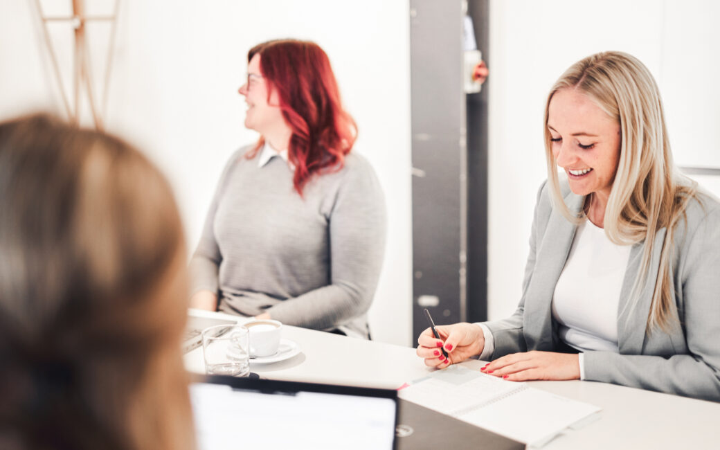 Drei Personen sitzen in einem hellen Raum an einem Tisch und unterhalten sich. Eine Person schreibt in ein Notizbuch, eine andere lächelt und die dritte ist teilweise zu sehen. Auf dem Tisch stehen Kaffeetassen und ein Laptop.