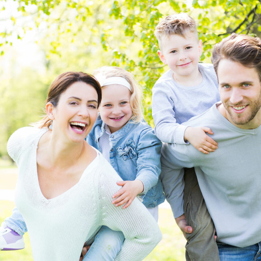 Bild von einer vierköpfigen Familie im Wald: Mutter und Vater tragen die Kinder huckepack und alle lachen in die Kamera.