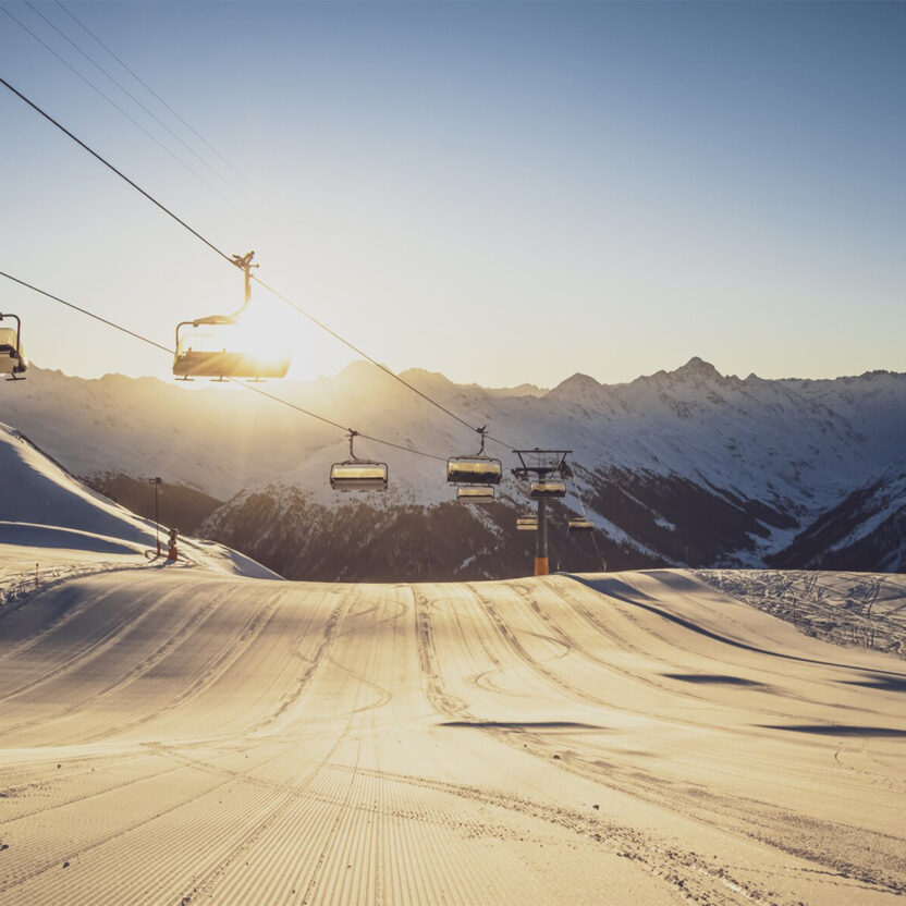 Davos Klosters Mountains Header: Zu sehen ist ein Sessellift, der im Gegenlicht über einer frisch präparierten Piste fährt.