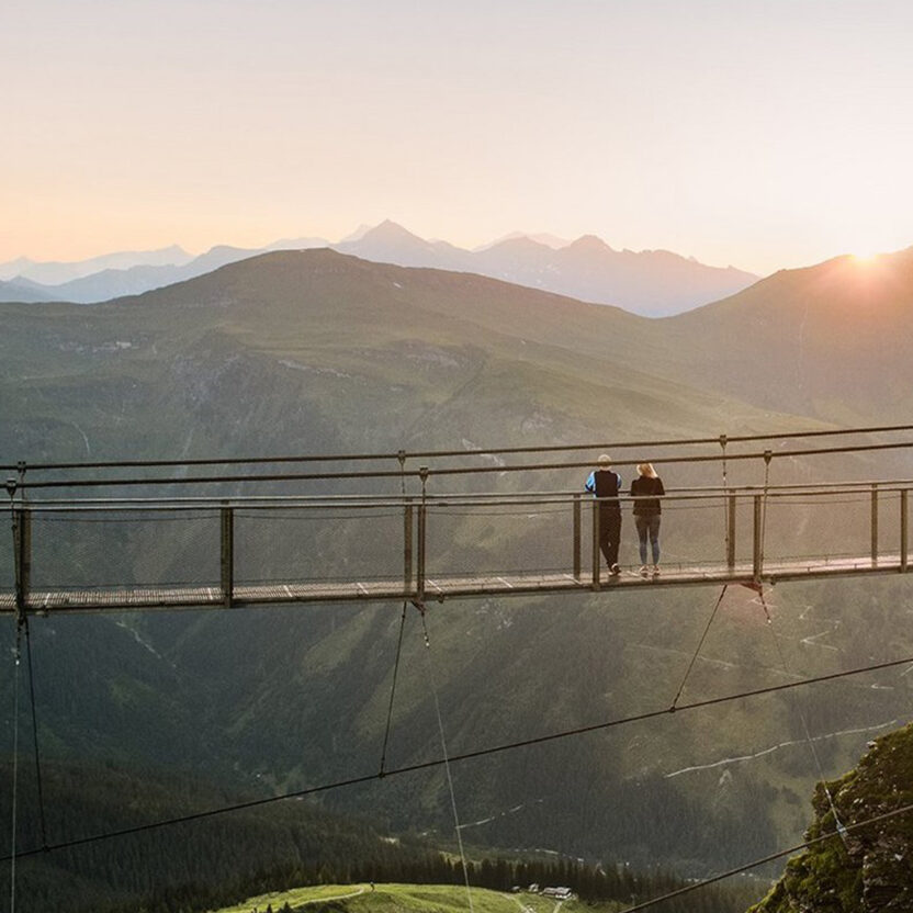 Bild von einer Hängebrücken in den Bergen von Skigastein, auf der zwei Personen stehen und in den Sonnenaufgang schauen.