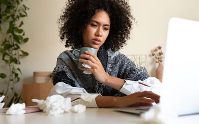 Sick African American woman working from home office using laptop drinking hot beverage covered with blanket. Sickness and working concept.