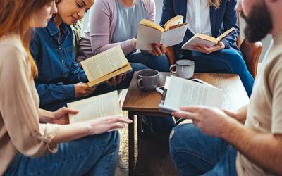 Small group of people with a mixed age range sitting at a table, discussing and reading books together.