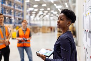 Supervisor standing by a whiteboard with a digital tablet discussing dispatch plan with workers. Team of workers having meeting in a distribution warehouse.