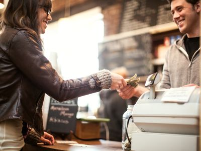 A happy woman hands a smiling cashier a few dollars at a coffee shop.