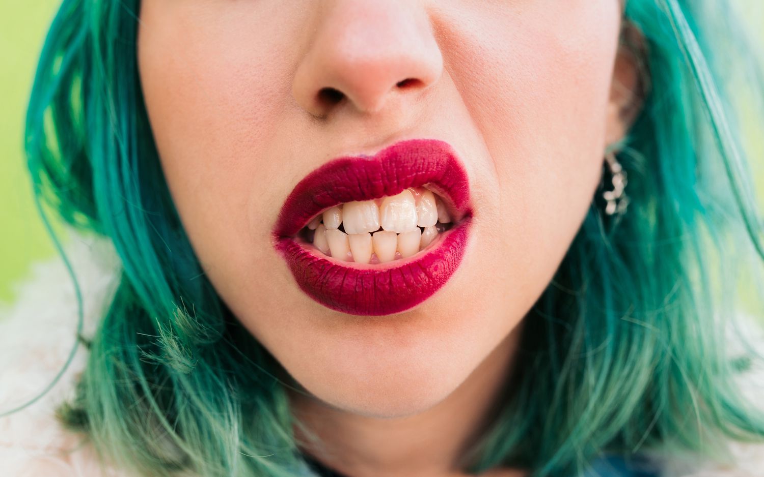 Detail of the mouth of a young woman with green hair and red lips making an aggressive gesture and showing her teeth. 