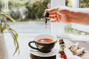 Hand dropping CBD oil into a Cup of Tea, surrounded by Cannabis Plants