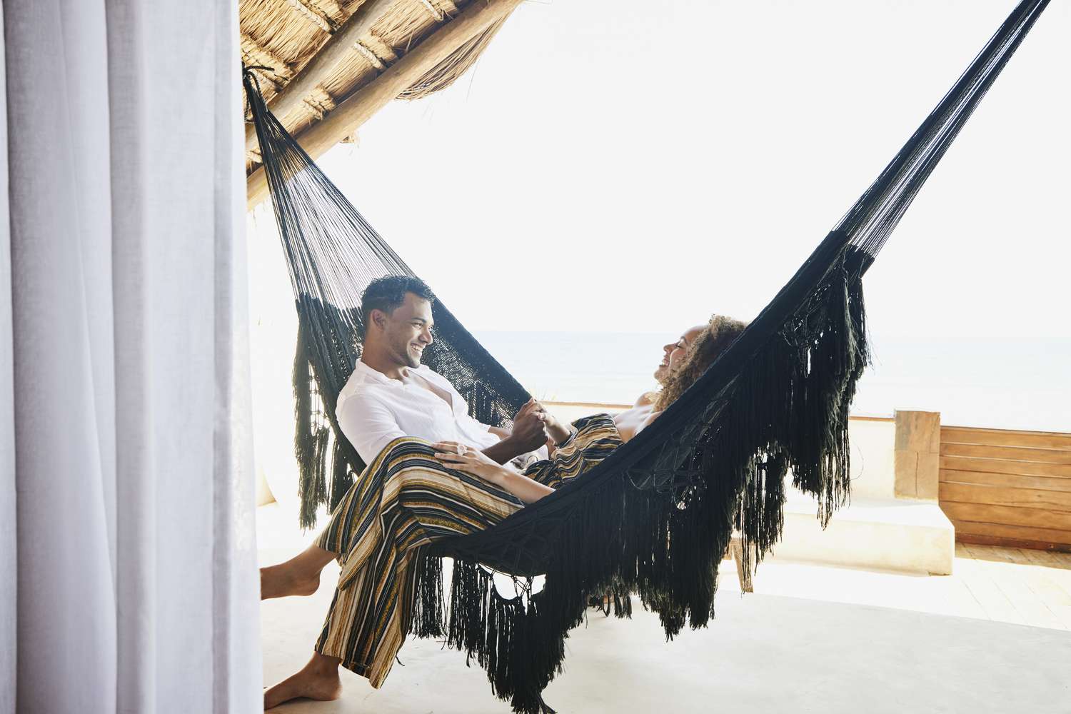 smiling couple holding hands while sitting in hammock on deck of luxury tropical villa