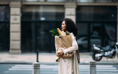 Woman with curly hair carrying a paper grocery bag crossing a street in an urban setting. She portrays a casual, everyday moment.