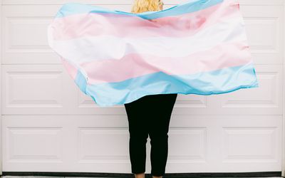 Transgender person from behind, wearing pink and white striped sweatshirt, holds transgender flag