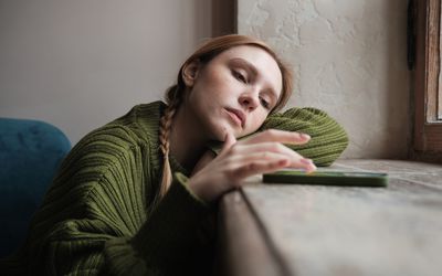 Upset sad millennial girl sitting by window looking at mobile phone waiting call from boyfriend