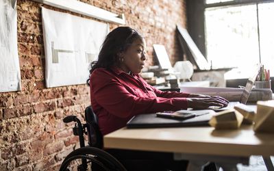 A woman seated in a wheelchair at a desk working on a laptop.
