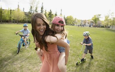 an extroverted mother giving her daughter a piggyback ride in sunny park