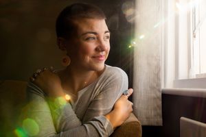 Young woman with short hair and a nose piercing hugs herself and looks out a window