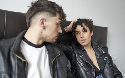 Man and woman having a serious conversation, sitting on a bedroom bed, dressed in casual clothes and staring at each other.