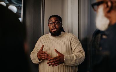 A man has a serious expression as he talks to onlookers. He uses his hands to gesture as he explains and discusses a matter with his coworkers / audience.