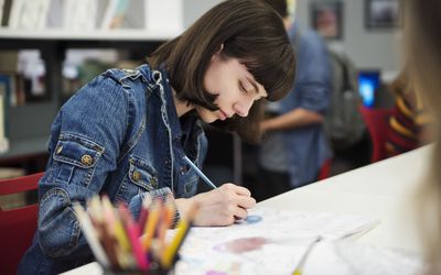 Young adult woman is colouring a mandala for relaxation in College library. Bookshelves and other students in the background. Horizontal waist up indoors shot with copy space.