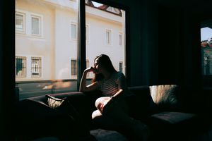 A tired and depressed Mediterranean woman is sitting on the sofa near the window at home, feeling exhausted and devastated. Emotional stress, burnout, and relationship difficulties that need to be taken care of
