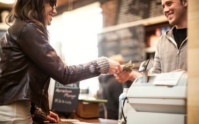 A happy woman hands a smiling cashier a few dollars at a coffee shop.