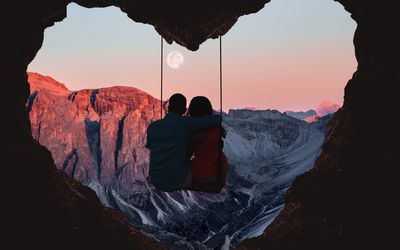 Couple on swing contemplating the mountains in a romantic view with heart shape. Composition of the Alps mountains during sunset with full moon and couple on swing from a heart shape cave.