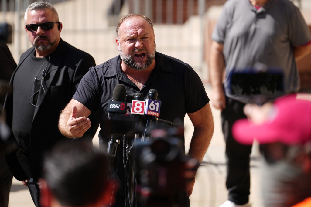 Alex Jones screams outdoors in front of a bank of microphones. He's wearing a black shirt and has a man who looks like private security behind him.