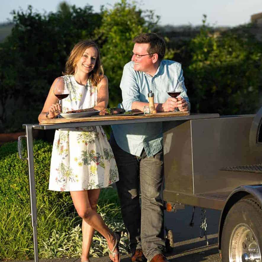 Sean Martin and Mary Cressler of Vindulge standing along side their catering BBQ trailer from Yoder.