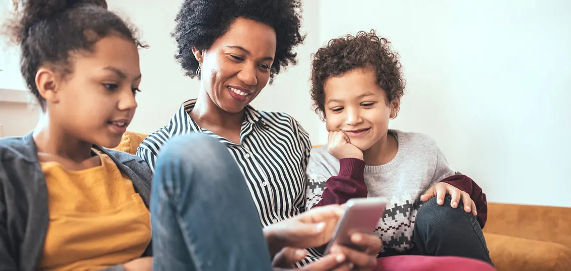 Shot of a mother sitting on the sofa with her kids looking at the mobile phone talking to older sibling at college